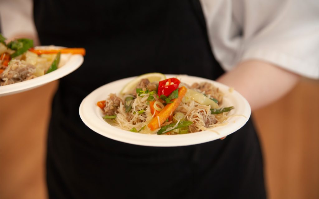 A person wearing an apron holds a plate of food featuring rice noodles with vegetables, sliced red chili, and meat. The dish includes carrots, green beans, and lime, presented on a white round plate.