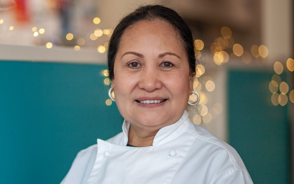 A smiling woman wearing a white chefs coat stands in front of a teal and white background with soft, blurred lights in the distance. She has long, dark hair pulled back and is wearing hoop earrings.