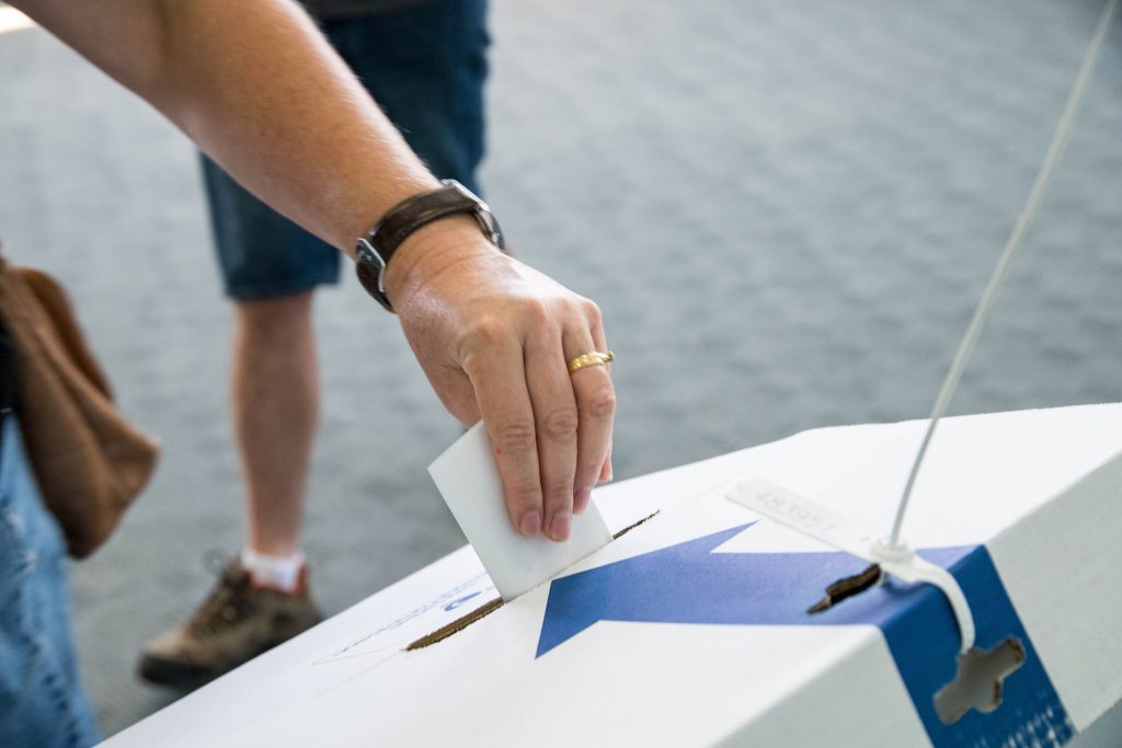 A person wearing a watch places a white paper into a voting box. The box is white with a blue star design. The background is blurred, showing another person standing nearby.