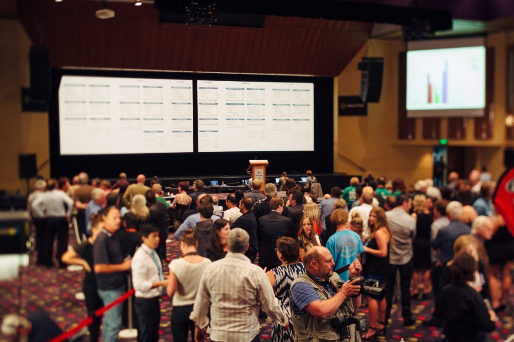 A large group of people gathered in an auditorium, facing a stage with two big screens displaying charts and data. A lectern is centered on the stage. Attendees are engaged in conversation.