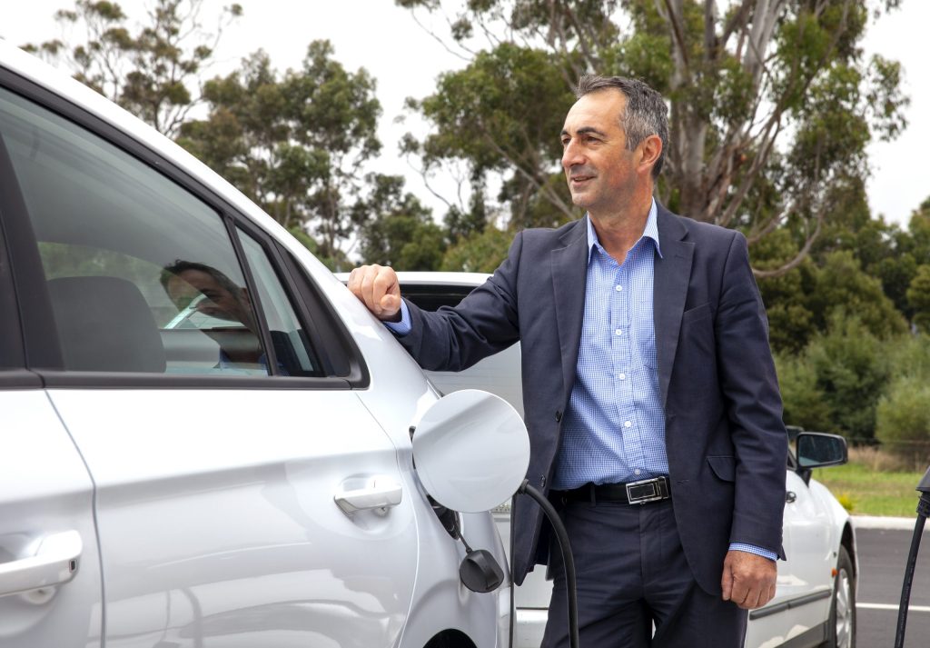 A man in a suit stands outdoors next to a white electric car, holding onto the open drivers door. The car is plugged into a charging station. Trees and a clear sky are in the background.