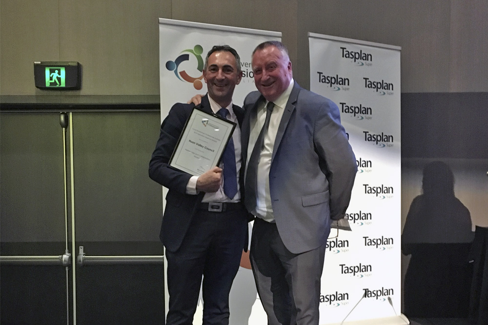 Two men in suits stand smiling indoors. One man holds a framed certificate. They are in front of a banner with logos and text, including Tasplan.