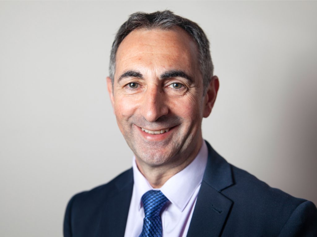 A man in a suit and tie smiles at the camera against a plain background.