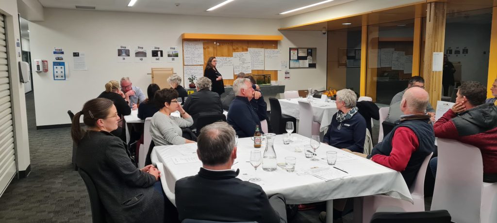 A group of people seated around tables listening to a woman speaking at the front of a room. The tables have white tablecloths and scattered papers. The room has bright lighting, with wooden and glass elements on one side.