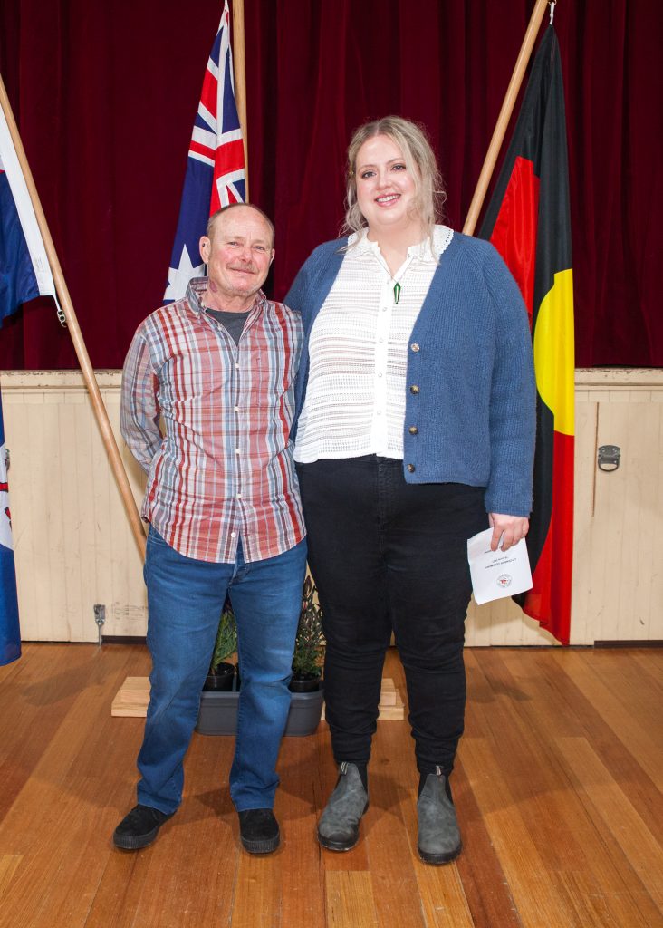 Two people stand smiling in front of four flags. The person on the left wears a plaid shirt and jeans, while the person on the right wears a blue cardigan over a white shirt and black pants. They are on a wooden floor with a red curtain backdrop.