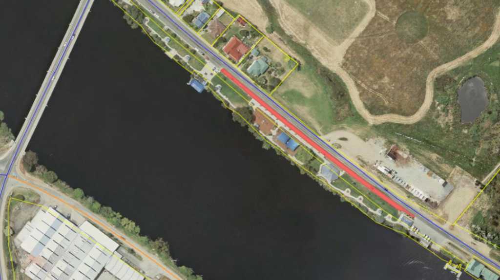 A satellite view of a road running alongside a water body. Houses are lined on one side, bordered by yellow lines, with fields and a small pond nearby. A bridge crosses the water at the bottom left, and industrial buildings are visible.
