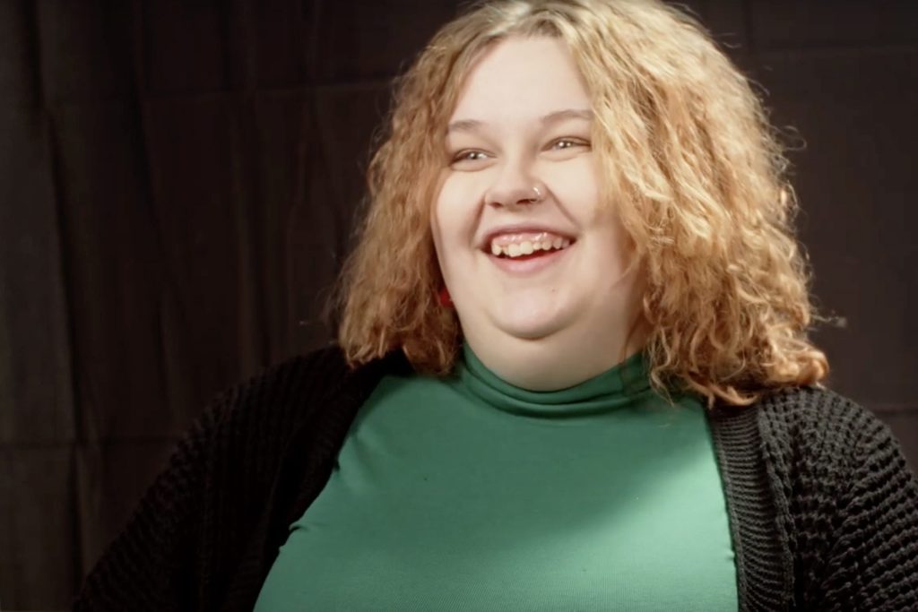 A person with curly hair and a nose piercing is smiling, wearing a green top and black cardigan, against a dark background.