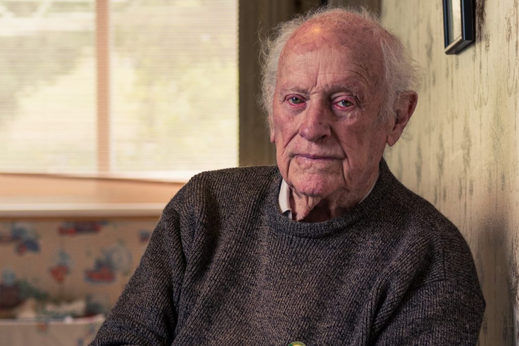 Elderly man with white hair and a thoughtful expression, wearing a dark sweater, seated indoors. Soft sunlight filters through a window, softly illuminating the room with patterned wallpaper and a wall with framed pictures.