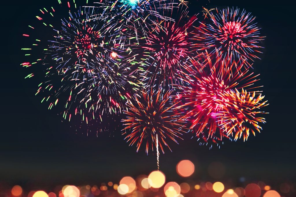 A vibrant display of fireworks bursts in the night sky, featuring multicolored explosions of red, blue, and white. The foreground is blurred with warm, glowing bokeh lights, adding depth to the scene.