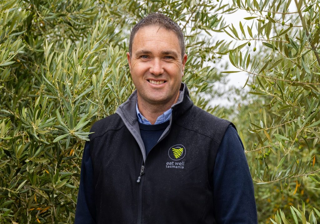 A man in a black vest with Eat Well Tasmania logo stands smiling among lush green olive trees. He is wearing a blue shirt and has short hair. The background shows dense foliage and a cloudy sky.