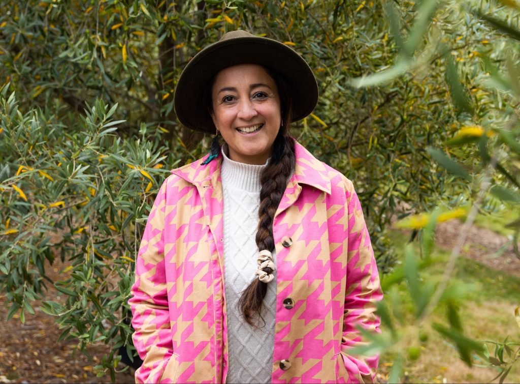 A woman stands smiling outdoors among green foliage. She wears a brown hat, a pink and yellow houndstooth coat, and a white sweater. Her dark hair is in a single braid. The setting appears to be a garden or park.