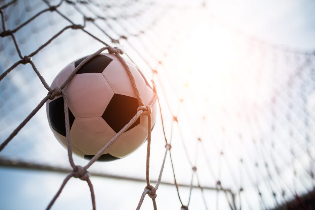 A soccer ball is captured in the net of a goal with sunlight streaming in from the top right corner, creating a bright, warm glow around the scene.