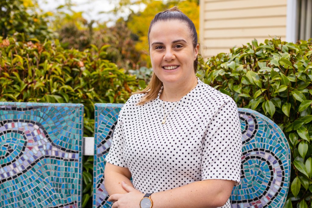 Woman smiling with her arms crossed in front of a colorful mosaic wall outdoors. She is wearing a white polka-dotted shirt and has her hair in a ponytail. Lush green foliage and part of a building are in the background.