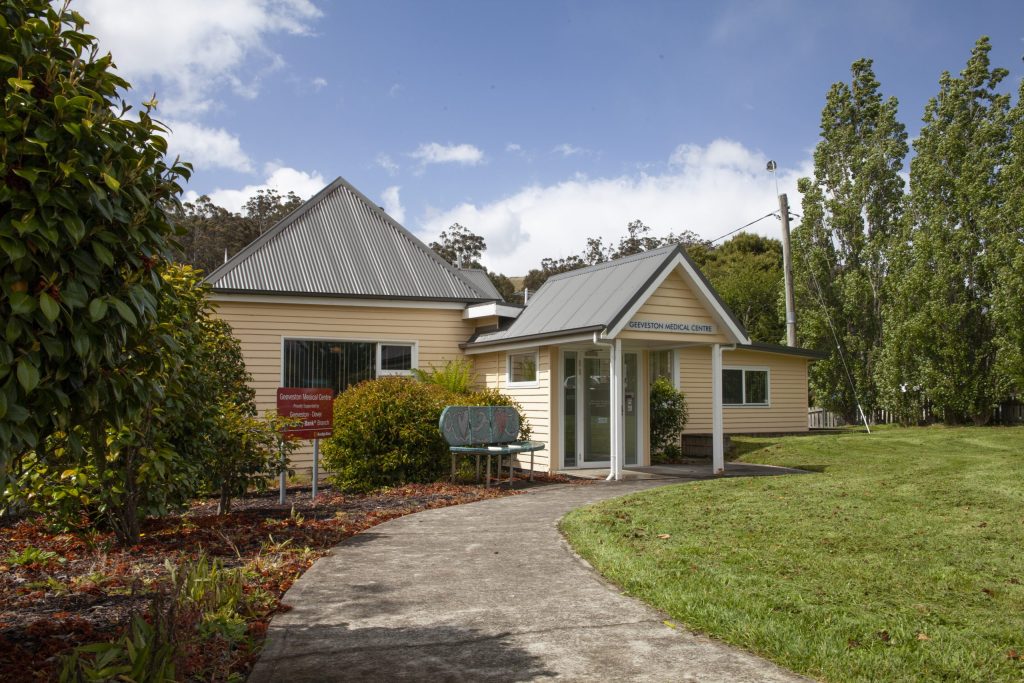 A small, yellow medical center with a metal roof and a glass entrance is surrounded by greenery and trees. A narrow, curved path leads to the entrance, and a red sign is visible on the lawn near the building. The sky is partly cloudy.
