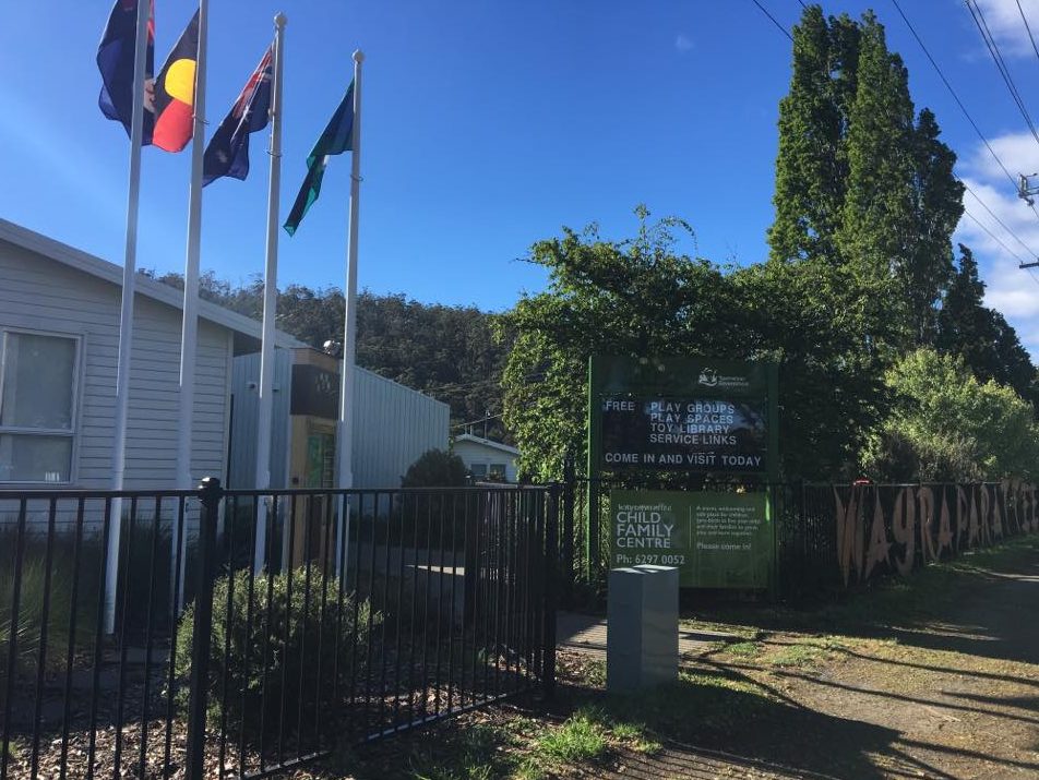 A community center with three flags on poles, including the Australian Aboriginal flag. A sign reads Child & Family Centre, and invites people to visit. Trees and a fence surround the building.