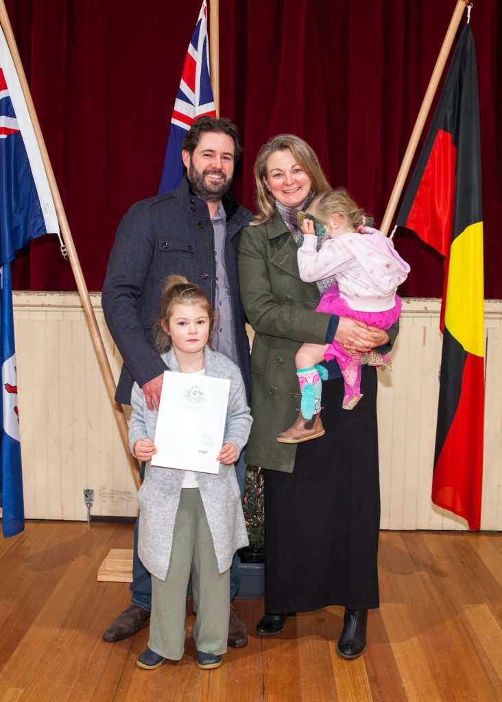 A family of four poses in front of two flags indoors. The woman holds a toddler, while the man stands beside a child holding a certificate. The background features a maroon curtain and wooden floor.