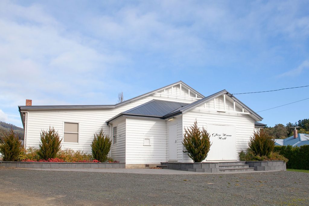 A white, single-story community hall with a gabled roof and sign saying Glen Huon Hall. It has multiple entrances, small windows, and is surrounded by shrubs and a gravel area. The sky is cloudy.