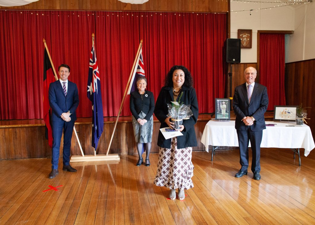 A woman stands holding a bouquet and certificate, surrounded by three people in a room with red curtains and flags. Two tables with framed pictures are in the background.