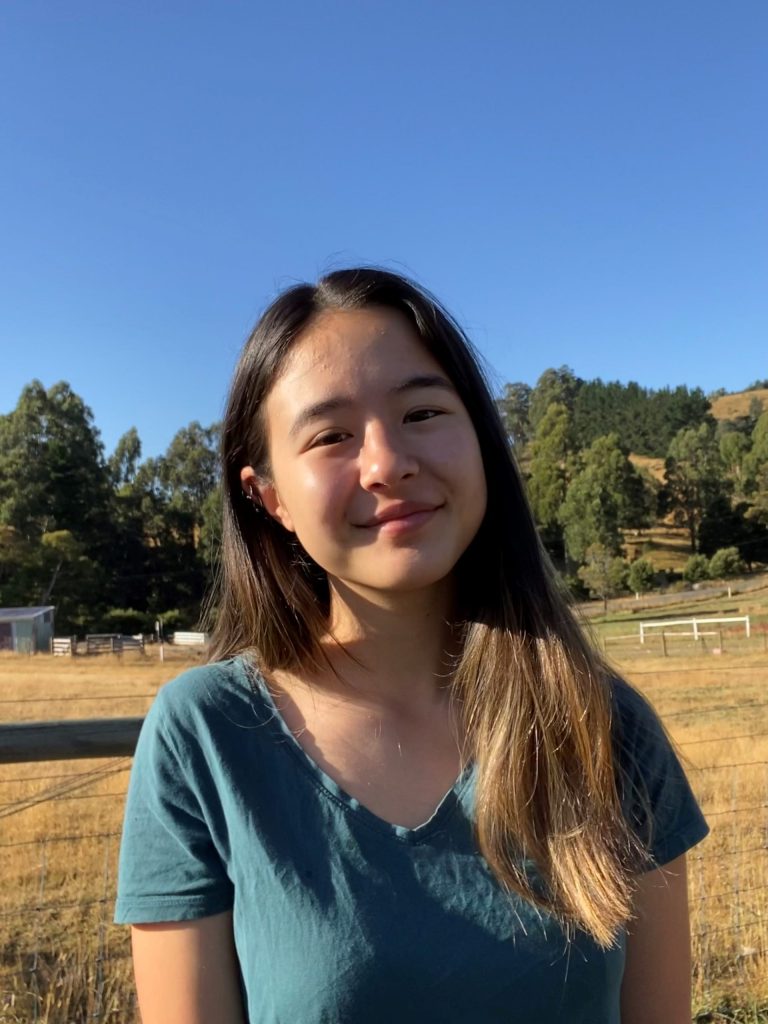 A young person with long dark hair stands outdoors in a sunny field, smiling. They wear a teal top. The background features grass, trees, and a clear blue sky.