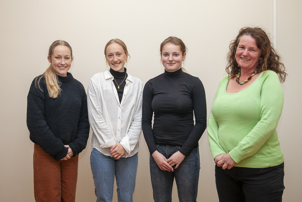 Four people stand indoors smiling at the camera. The three individuals on the left wear casual sweaters and jeans, while the person on the right is in a green top with a necklace. They are in front of a plain beige wall.