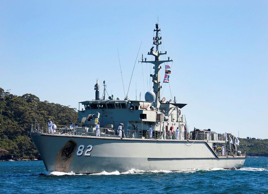 A naval ship with the number 82 on its side sails through clear blue water. Several crew members are visible on deck, and a line of signal flags is strung along the bridge. A forested shoreline is in the background under a clear sky.