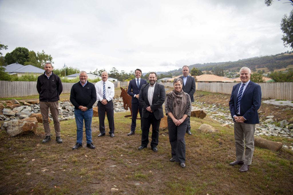 A group of nine people in formal and casual attire stand on a grassy area with rocks and plants. A wooden sculpture is visible behind them. Trees and suburban houses are in the background under a cloudy sky.