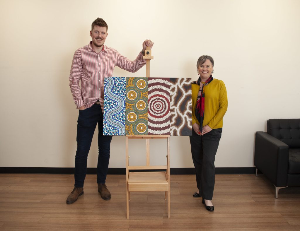 A man and a woman stand on either side of a colorful piece of abstract art on an easel. The artwork features intricate patterns in blue, green, gold, and maroon. They are in a room with a wooden floor and a dark couch.