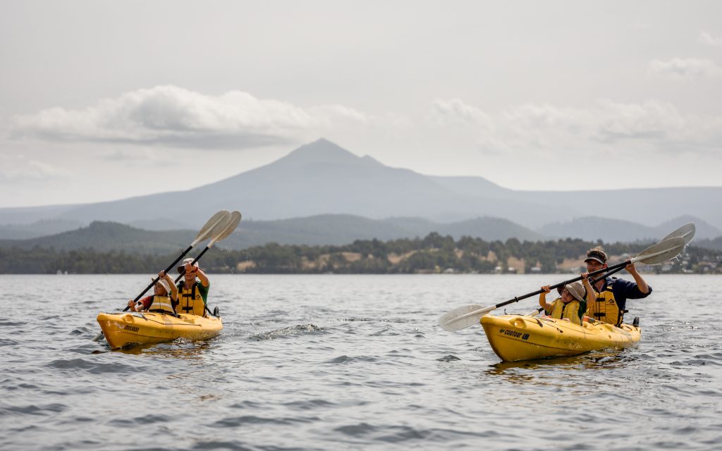 Two people kayaking on a calm lake, each in a yellow kayak. They are paddling toward the camera, with a mountain and cloudy sky visible in the background. The scene conveys a sense of adventure and tranquility.