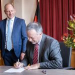 Two men in suits; one stands smiling, the other sits signing a document. The setting includes red curtains, a floral arrangement, and flags in the background, suggesting an official event or agreement.