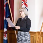 An older woman with gray hair reads a document while standing in front of three flags. She is wearing glasses, a black top, and a patterned skirt in a room with red curtains and wooden paneling.