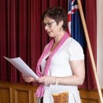 A woman in a white shirt and pink scarf reads from a paper. She stands with curtains and two flags in the background.
