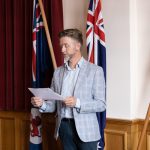 A man in a blue plaid blazer is speaking while holding a piece of paper. He stands in front of two flags in a room with wooden paneling and maroon curtains.