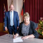 A woman is sitting at a table, signing a document, while a man in a suit stands beside her, smiling. Behind them are red curtains and two flags. A vase with flowers is on the table.