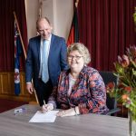 A woman sitting at a table and signing a document, smiling. A man stands beside her, also smiling. Behind them are red curtains and flags. A floral arrangement is on the table. The setting appears formal.