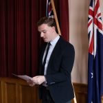 A person in a suit reads from a paper, standing in a room with red curtains. Two flags featuring the Union Jack and Southern Cross are displayed behind.