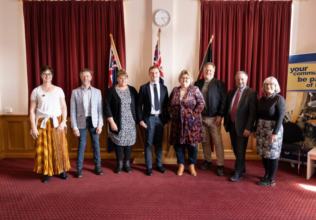 A group of eight people stands in a row in a room with red curtains and two flags. They are dressed in business and casual attire. A clock is on the wall above them.