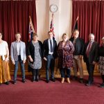 A group of eight people stands in a row in a room with red curtains and two flags. They are dressed in business and casual attire. A clock is on the wall above them.