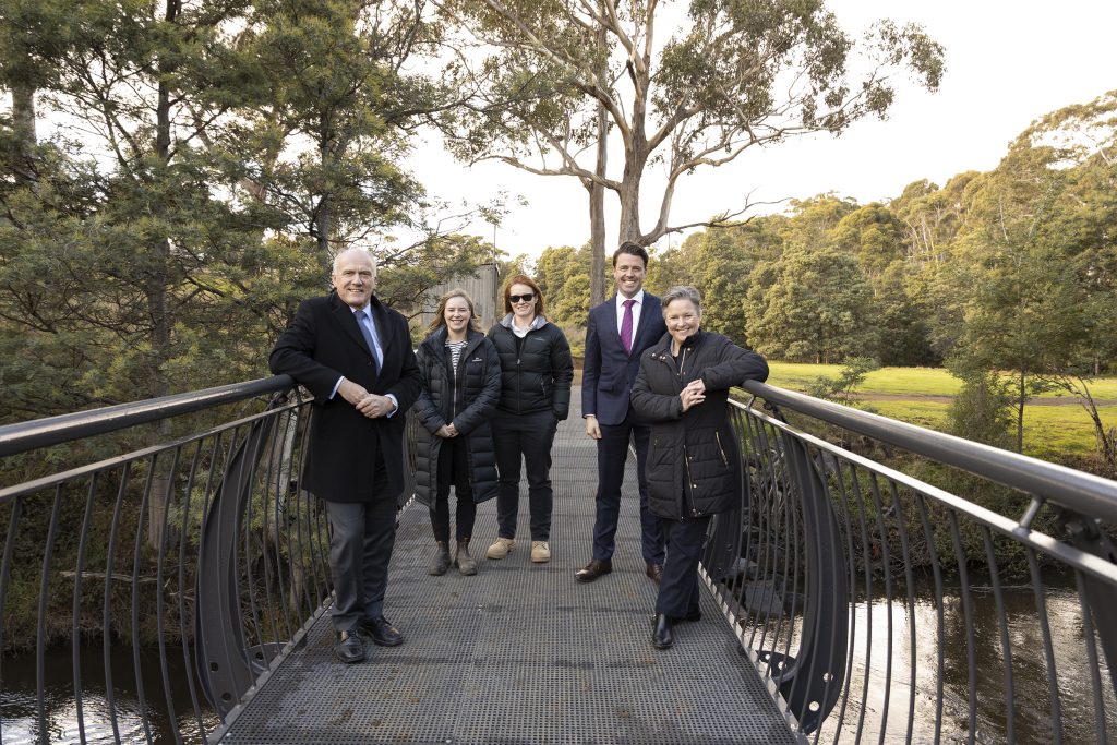 A group of five people, wearing formal and casual attire, stand on a metal footbridge in a park. Trees and greenery surround them under a clear sky. They pose for the camera, smiling.