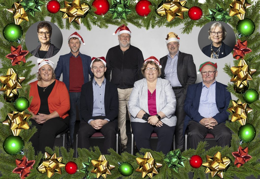 A group of eight people, some wearing Santa hats, arranged in two rows. They are framed by a festive border of green leaves and colorful ornaments. Four circular inset photos of individuals are in the corners of the frame.