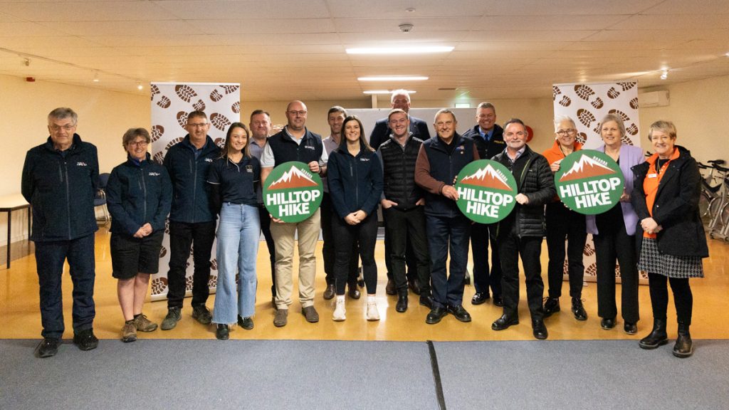 A group of people standing indoors, smiling and holding signs that say Hilltop Hike. They are in front of banners with a logo. The group is diverse, with men and women wearing jackets and casual clothing.