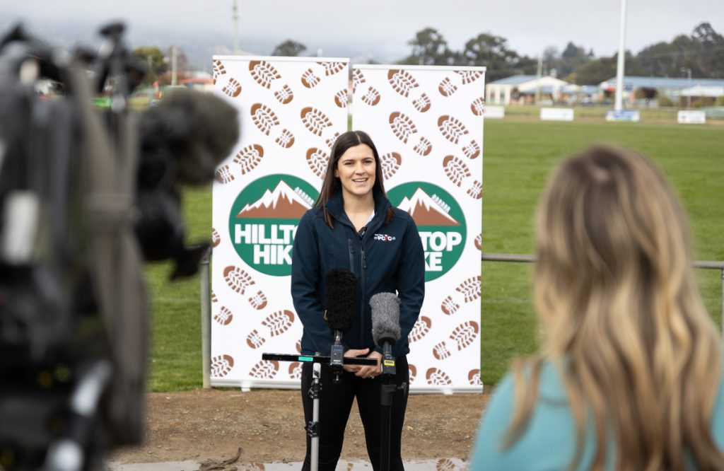 A woman stands in front of banners with Hilltop Hike logos, speaking at a microphone. She is outdoors on a field, likely giving an interview or speech. Camera equipment and another person are visible in the foreground.