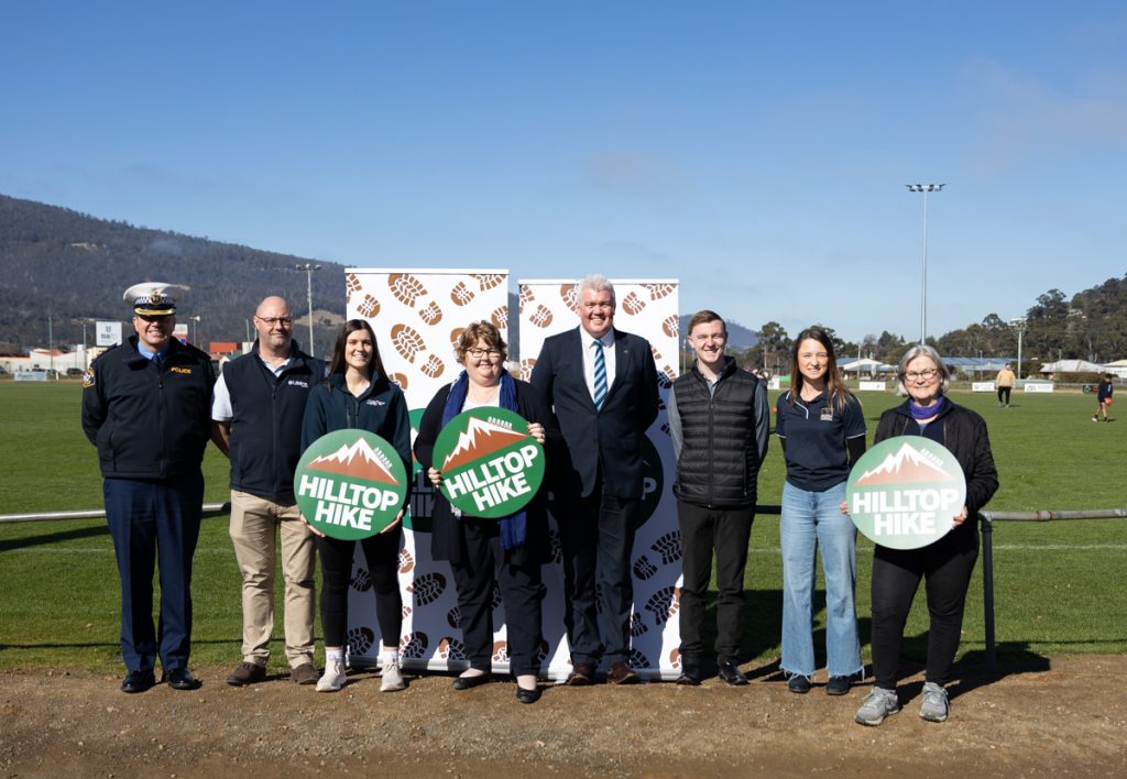 A group of eight people stand outdoors on a grassy field, posing for a photo. Two individuals in the front hold Hilltop Hike signs. A backdrop with logos is behind them, and a clear sky and hills are in the distance.