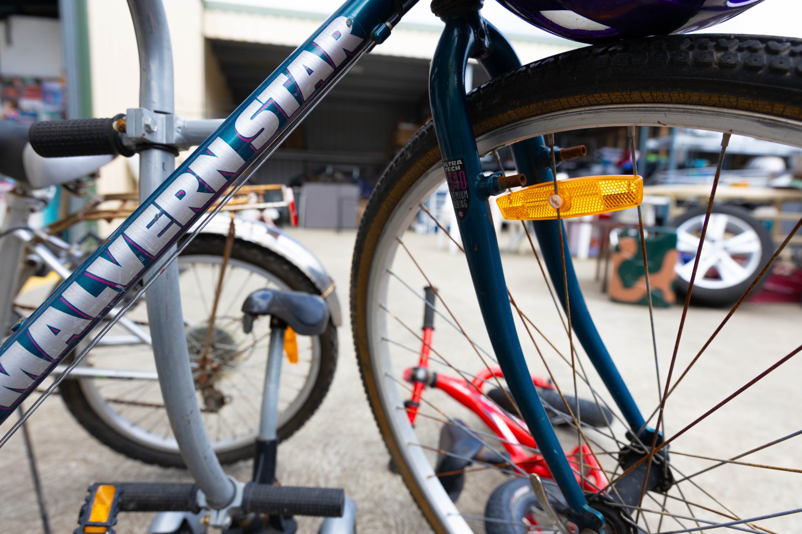 Close-up of a blue bicycle frame with MALVERN STAR written on it. The focus is on the wheel and reflector. A red bicycle lies on the ground in the background inside a garage or storage area.