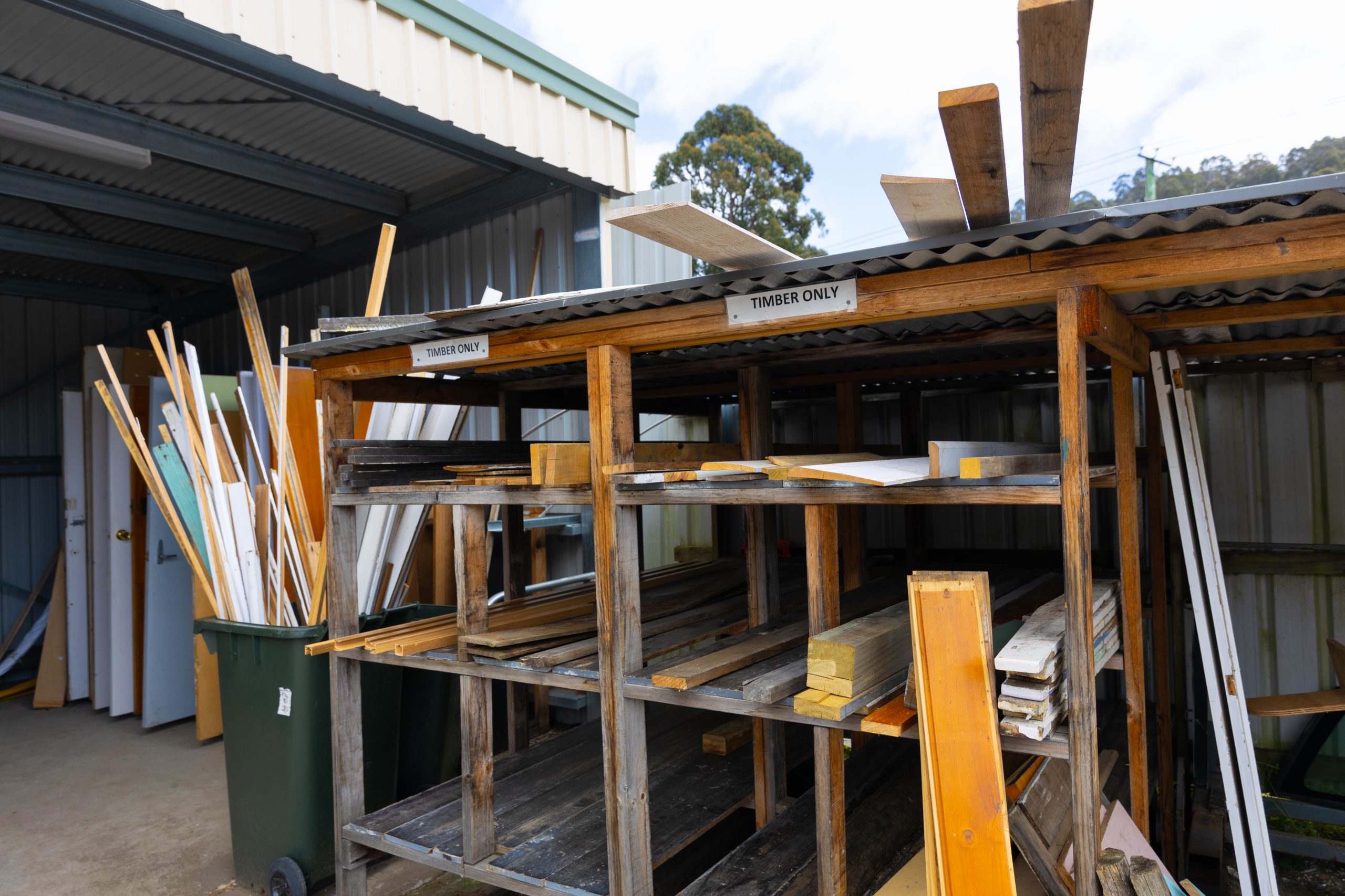 Wood storage area with assorted planks and beams organized on metal racks. Signs labeled Timber Only are attached. The setting appears to be an outdoor shed or workshop with a corrugated metal roof and walls.