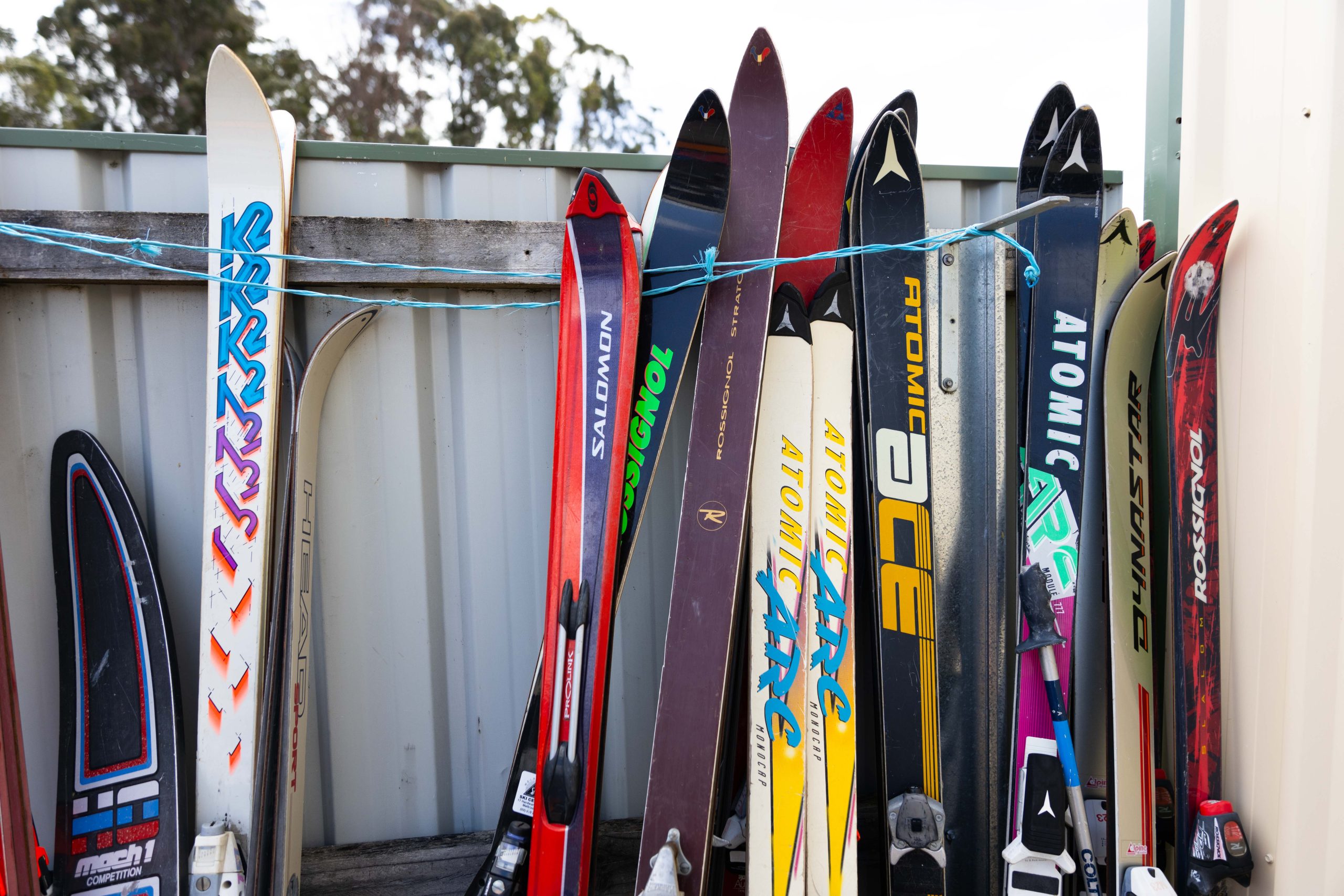 A row of colorful skis and ski poles lined up against a metal wall. Various brands and designs are visible, with some skis standing upright and others leaning. Blue cord is strung across for additional support. Trees are visible in the background.