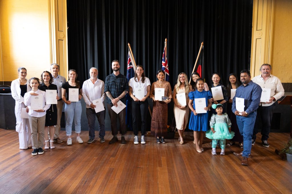 A diverse group of people stand on a wooden stage holding certificates, smiling. Behind them are three flags. The setting appears to be an indoor auditorium with black curtains.