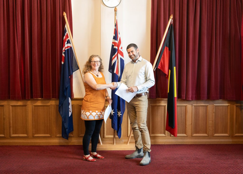 A woman in an orange dress and a man in a white shirt shake hands in front of three flags. They stand in a room with red curtains and wooden paneling, holding a document between them.