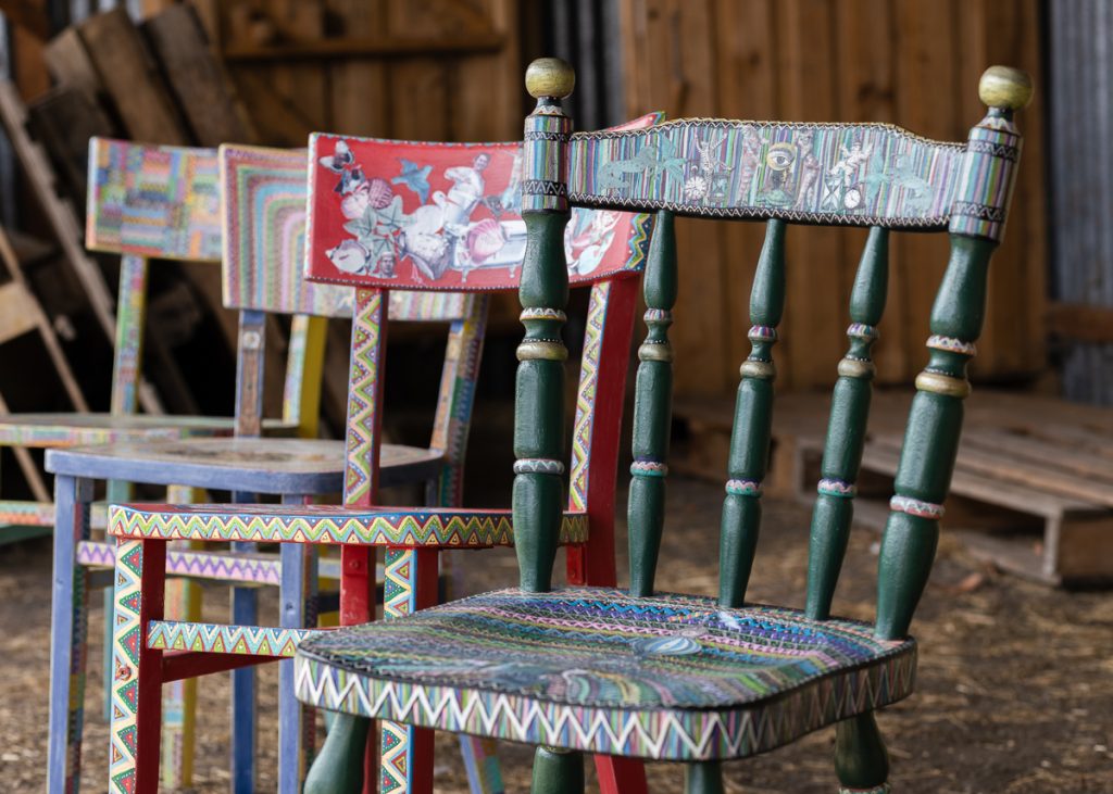 Three intricately painted wooden chairs in a rustic setting. The chair in the foreground is green and decorated with detailed patterns. The chairs behind feature diverse colorful designs, adding a vibrant touch to the wooden backdrop.