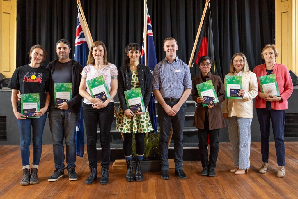 Seven people standing in a row, each holding a green certificate. There are two flags behind them on a stage with dark curtains. The setting appears to be a formal event or ceremony.