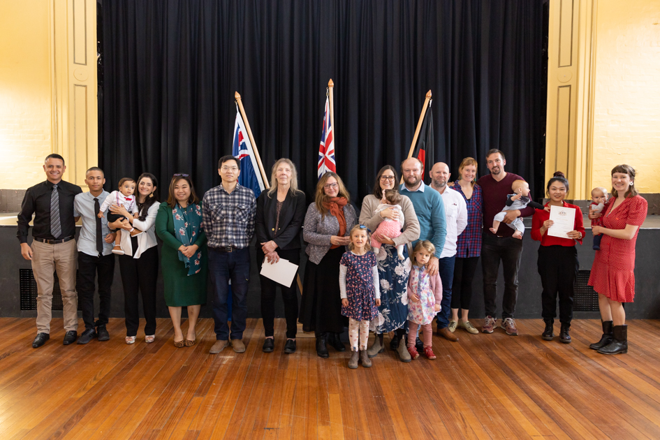 A diverse group of adults and children stand indoors on a wooden floor, facing the camera. Three flags are displayed in the background against a dark curtain. Some adults hold babies, and one person holds a certificate. Everyone appears cheerful.